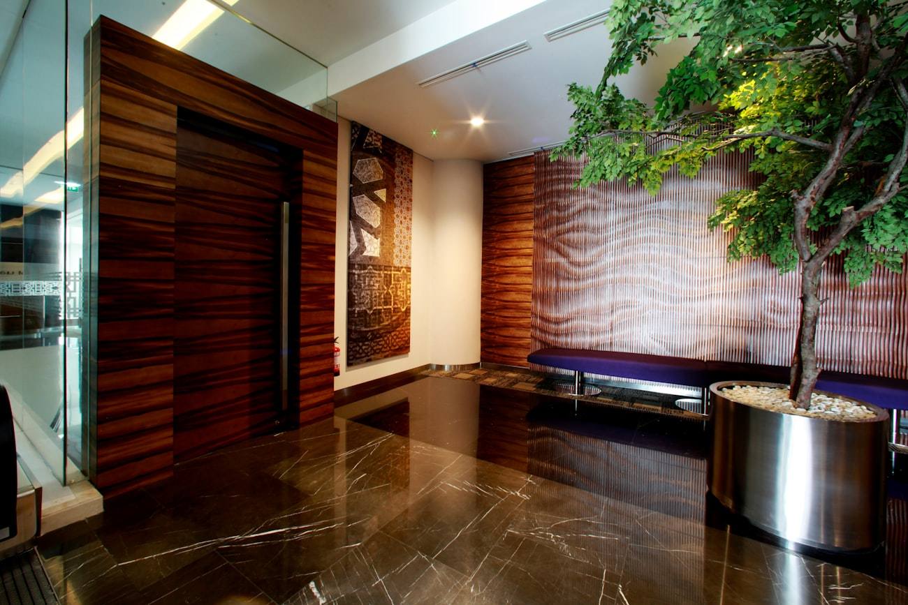 Portal de acceso con revestimiento de madera, butacas y un árbol interior.